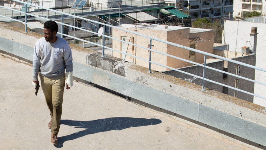 John David Washington on a Greek rooftop holding a gun as seen on the official poster for the new Netflix thriller BECKETT.
