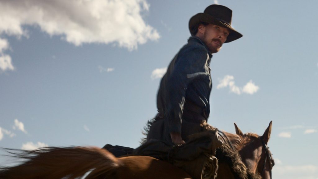 Benedict Cumberbatch wearing cowboy gear as he rides a brown horse against a blue cloudy sky as seen in THE POWER OF THE DOG, premiering at the 2021 Venice film festival and coming soon to Netflix.