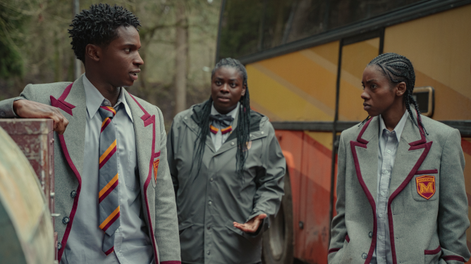 Jackson, Viv, and Cal stand together in front of a school bus all wearing the new Moordale school uniforms as seen in SEX EDUCATION season 3 on Netflix.