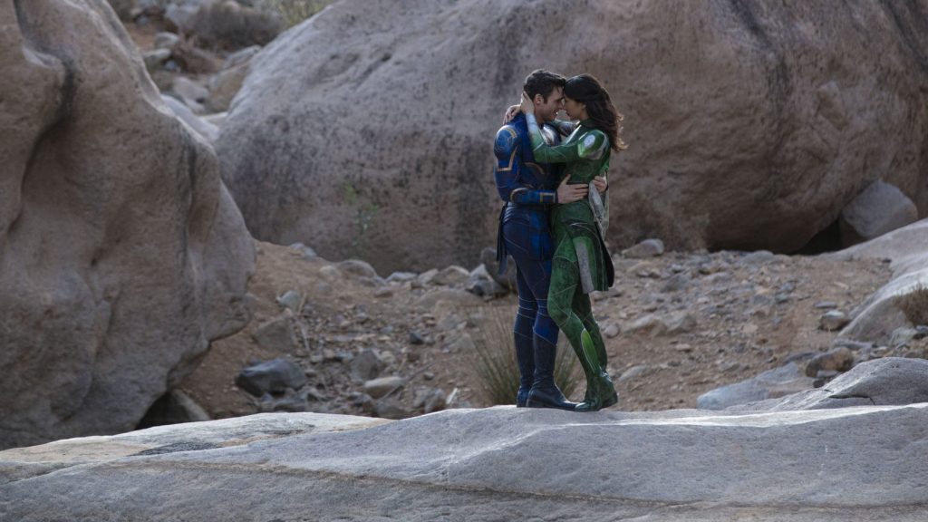 Richard Madden as Ikaris and Gemma Chan as Sersi embrace each other passionately while wearing their trademark blue and green cosmic suits in front of a rock formation as seen in the new Marvel Studios film ETERNALS directed by Chloé Zhao.