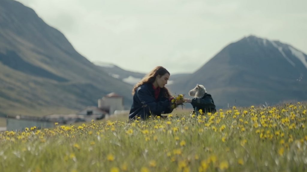 Noomi Rapace collects yellow flowers with her new lamb hybrid child Ada on the Icelandic hillside with snowy mountains in the backdrop as seen in the new A24 film LAMB.