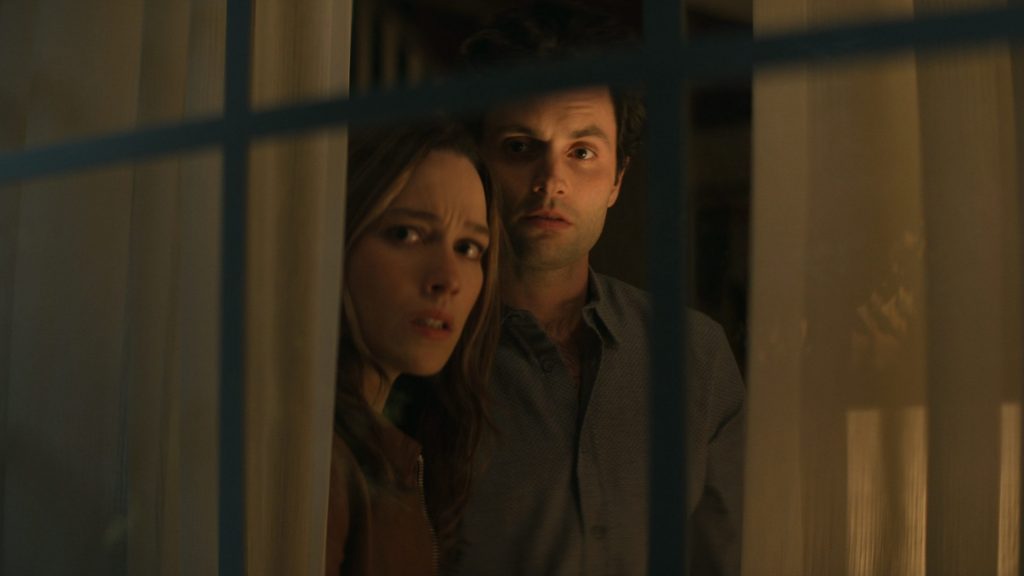 Penn Badgley as Joe Goldberg and Victoria Pedretti as Love Quinn peak through the blinds of their new suburban home in shock as seen in season 3 of the Netflix original thriller series YOU.