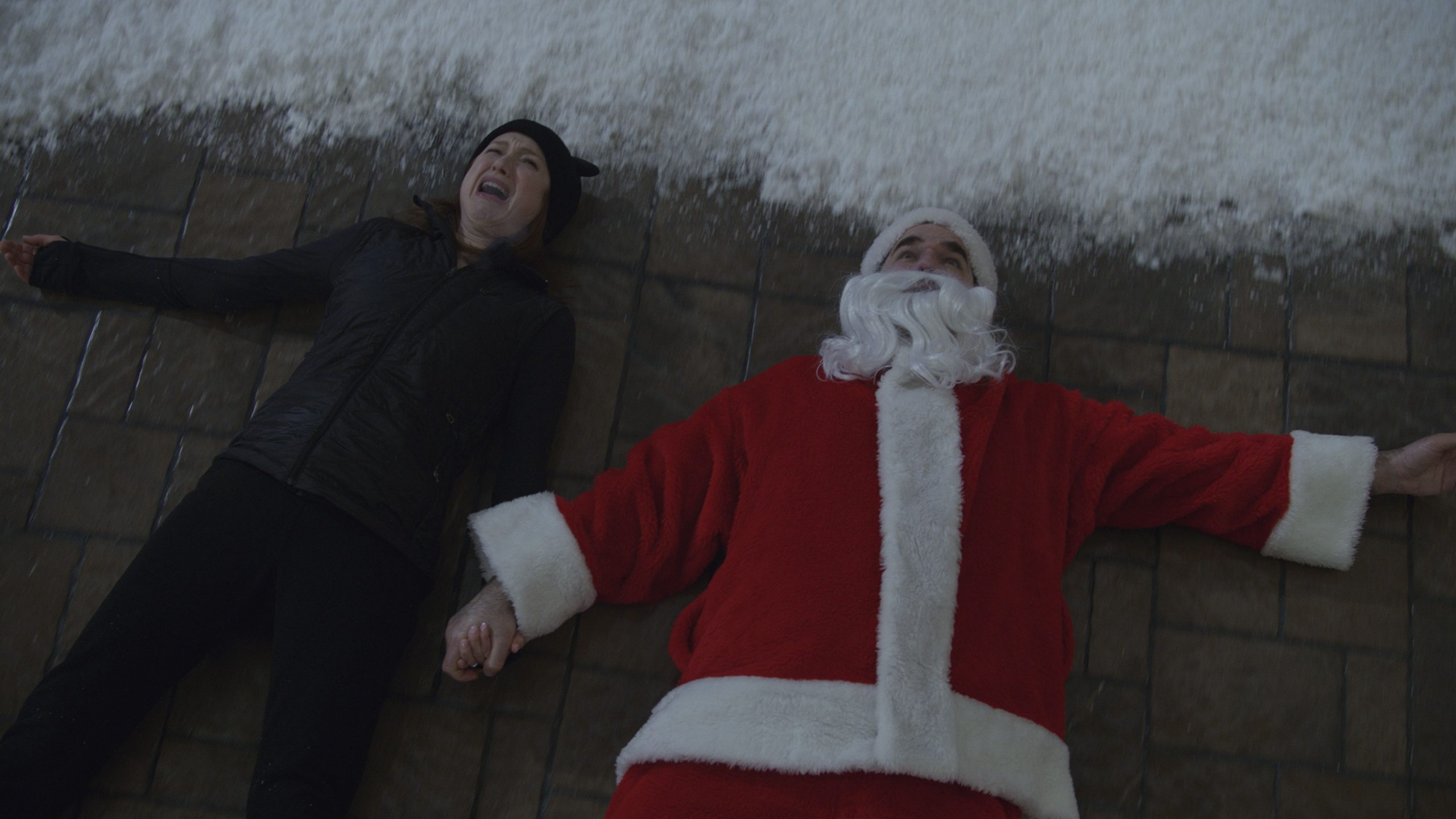 Ellie Kemper and Rob Delaney dressed as a burglar and Santa Claus as they hold hands on the sidewalk floor in pain as seen in the new Home Alone sequel exclusive to Disney+ HOME SWEET HOME ALONE.