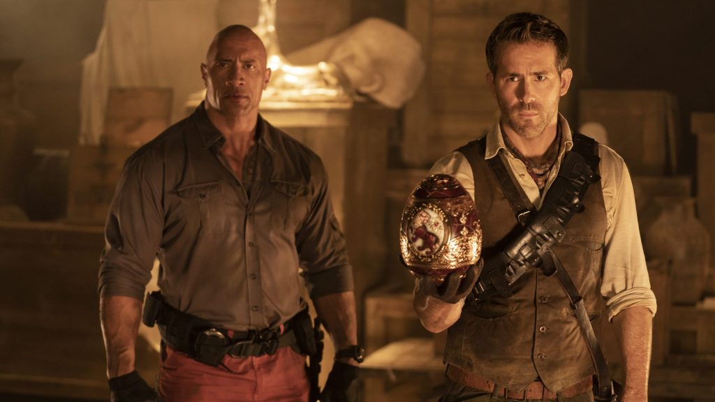 Dwayne Johnson and Ryan Reynolds stand side by side in jungle gear as Reynolds holds a rare golden Egyptian egg as seen in the original Netflix action comedy RED NOTICE.
