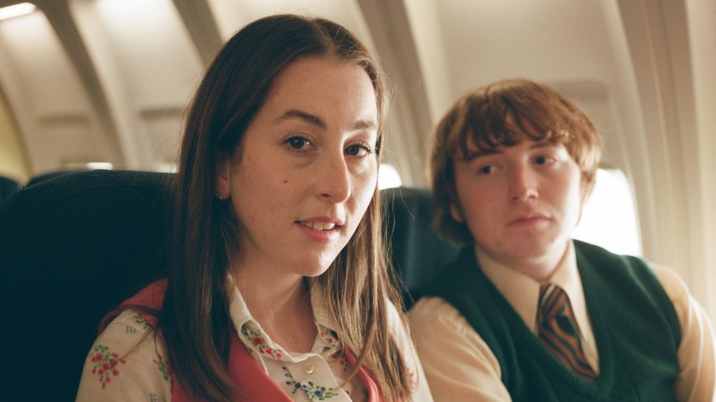 Alana Haim and Cooper Hoffman sit next to each other on a plane as Cooper stares at her with googly eyes and she looks into the camera as seen in LICORICE PIZZA directed and written by Paul Thomas Anderson.
