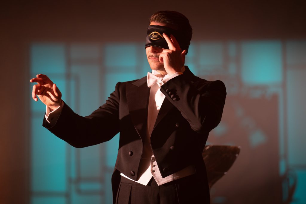 Bradley Cooper as Stanton Carlisle wearing a satin blindfold with a cyclops eye printed on as he waves his hand around with the other on his temple while doing a mentalist trick reading in NIGHTMARE ALLEY directed by Guillermo Del Toro.