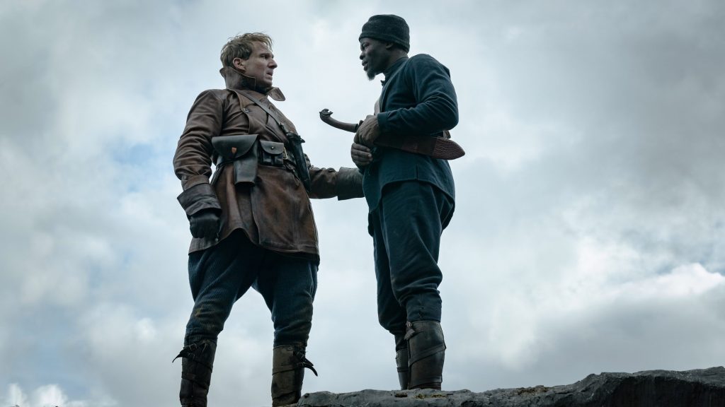 Ralph Fiennes as Oxford and Djimon Hounsou as Shola in their battle uniforms standing on the edge of a cliff as seen in THE KING'S MAN directed by Matthew Vaughn.