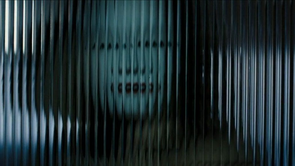 Rami Malek as the villainous Safin wearing a white mask behind a layered glass door as shot by cinematographer Linus Sandgren in NO TIME TO DIE. 