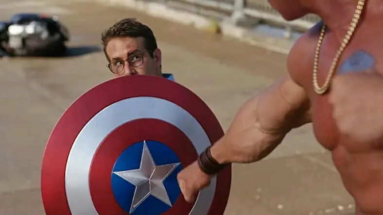 Ryan Reynolds protects himself with Captain America's shield while facing off against the bigger "Guy" that has a blue shirt tattoo on his chest in FREE GUY, coming to Disney+ in February 2022.