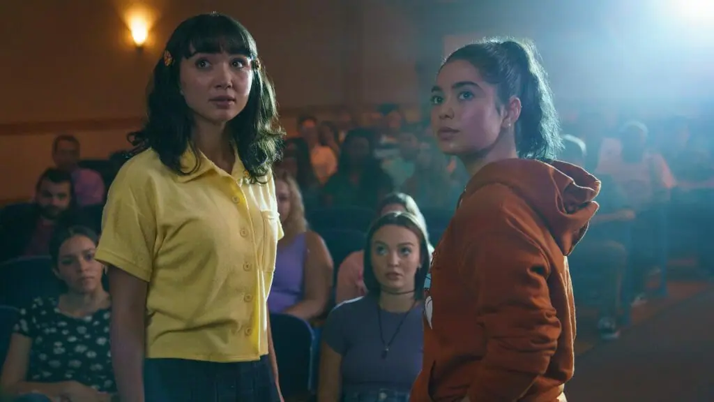 Rowan Blanchard and Auli'i Cravalho are struck by a theater spotlight as they both stand up in front of a sitting auditorium crowd in the new lesbian teen rom-com CRUSH on Hulu.