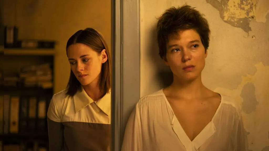 Kristen Stewart and Léa Seydoux stand and look distressed on the opposite sides of a door frame in an apartment with peeling walls as seen in CRIMES OF THE FUTURE.