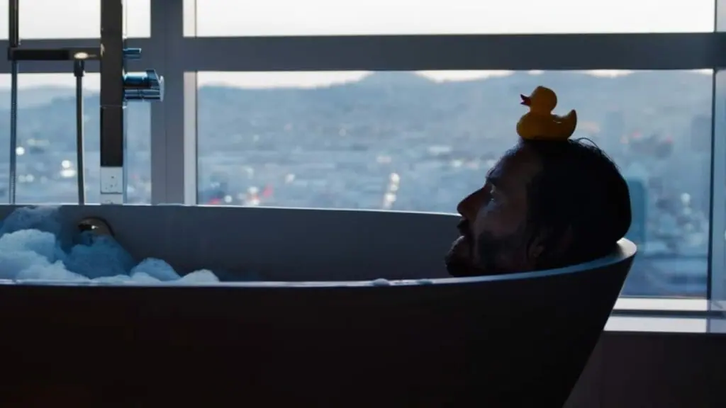Keanu Reeves as Neo sits quietly in a bath tub of bubbles with a rubber duck on top of his head in THE MATRIX RESURRECTIONS, new on HBO Max in May 2022.