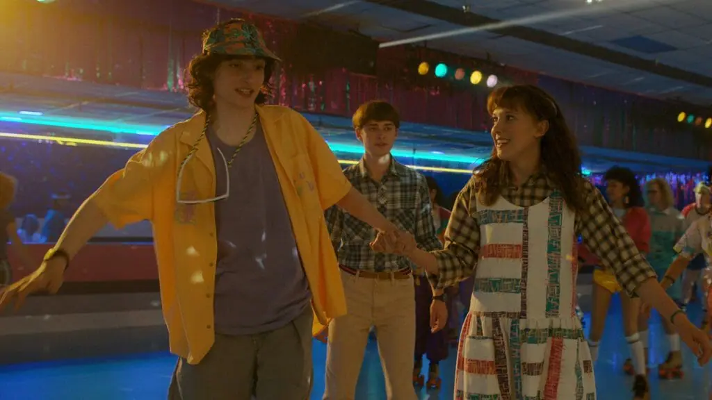 Eleven holds Mike Wheeler's hand and laughs as he struggles to skate in a neon bright 80s roller rink while Will Byers follows between them in season 4 of STRANGER THINGS on Netflix.