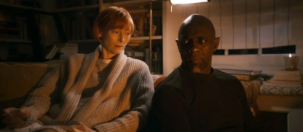 Tilda Swinton sits in a bath robe next to Idris Elba as a magical Djinn with pointy elf ears in a warm Istanbul hotel room as seen in THREE THOUSAND YEARS OF LONGING directed by George Miller.