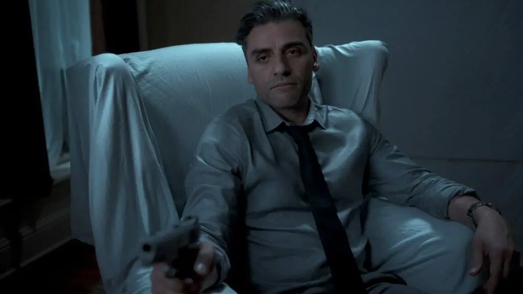 Oscar Isaac holds up a gun menacingly while sitting in a chair covered in white sheets in THE CARD COUNTER, new on HBO Max in June 2022.