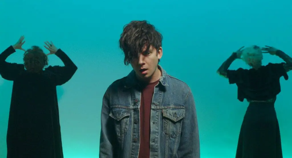 Asa Butterfield sports a jean jacket and large punk hairstyle that covers his right eye in front of a turquoise blue backdrop with two ladies posing with their hands up on the left and right as seen in FLUX GOURMET..