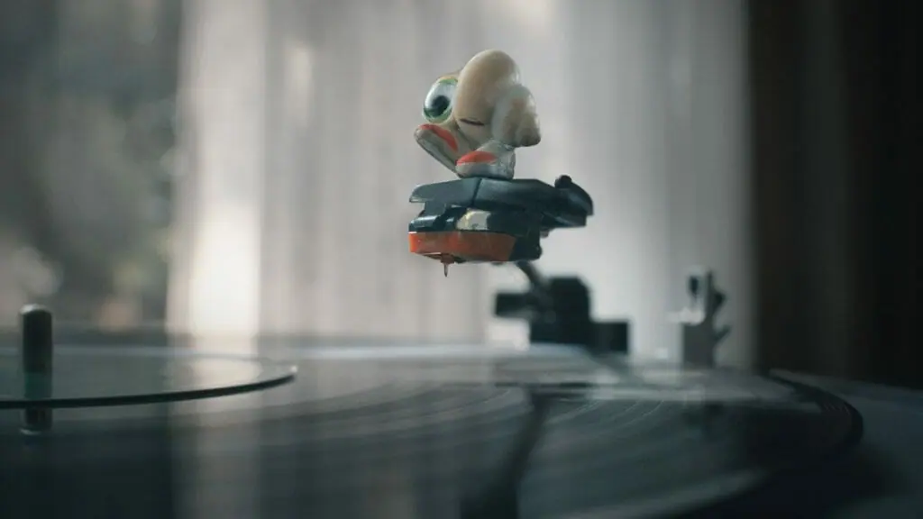 Marcel the Shell sits on top of a record player needle with his shoes dangling as a black vinyl record spins below him in the A24 stop motion animated film MARCEL THE SHELL WITH SHOES ON.