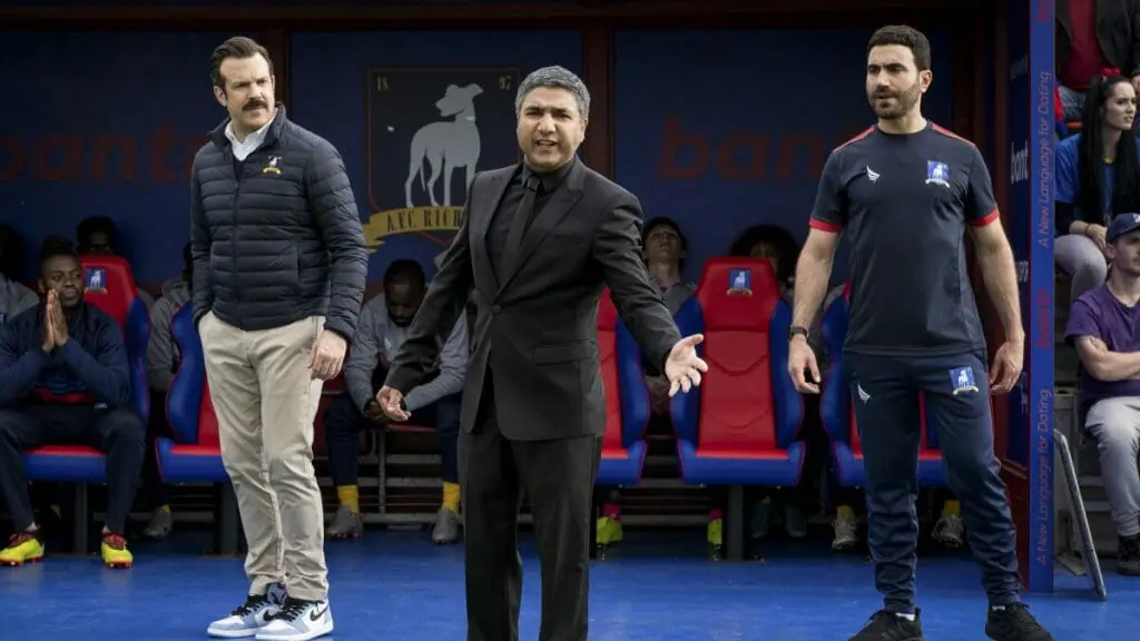 Nate played by Nick Mohammed in a black suit yells at the field as the coach of AFC Richmond during a game while Ted Lasso and Roy Kent watch concerned from behind in TED LASSO Season 2 on AppleTV+.