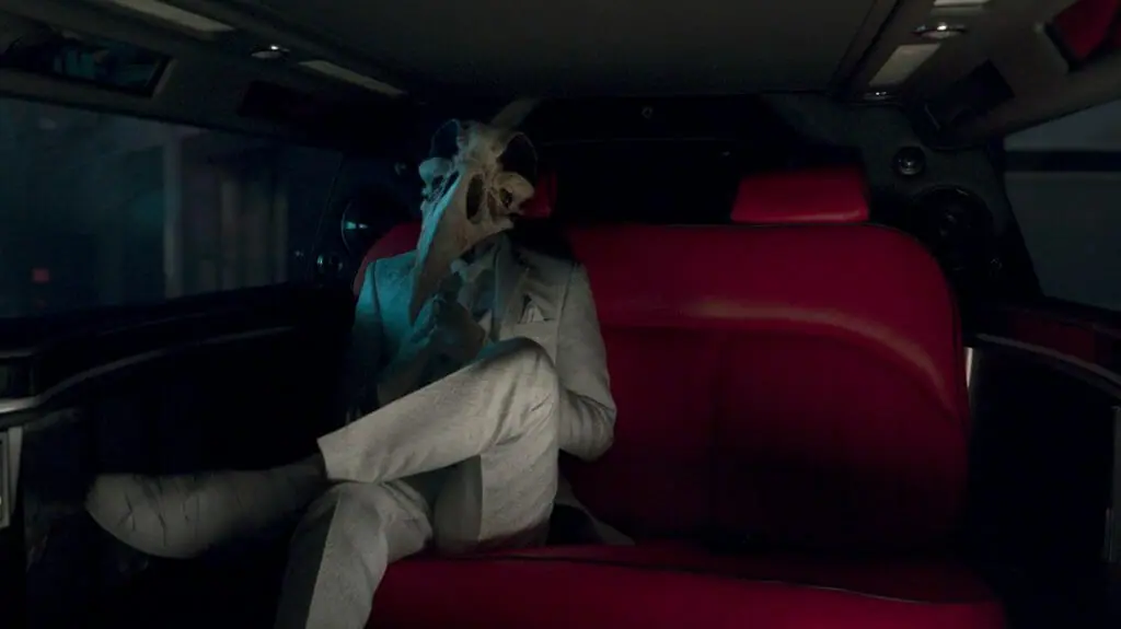 Khonshu voiced by F. Murray Abraham sits in the back of a dark limo wearing a clean white suit and tie with his legs crossed in the Disney+ MCU series MOON KNIGHT.
