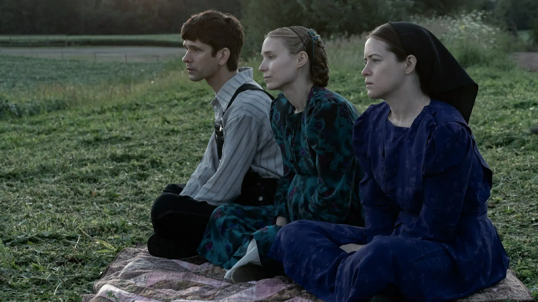 Ben Wishaw, Rooney Mara, and Claire Foy play members of an old Mennonite colony sitting together on a grassy hill in the drama film WOMEN TALKING.