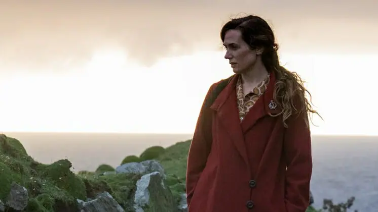 A cinematic shot of Kerry Condon wearing a burgundy red coat in front of a beautiful island sunset in THE BANSHEES OF INISHERIN.