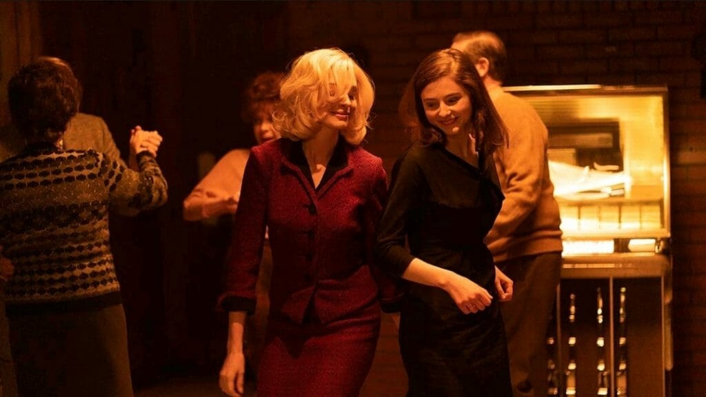 Anne Hathaway and Thomasin McKenzie dance closely together in a dimly lit bar in the period noir twisted romance film EILEEN.
