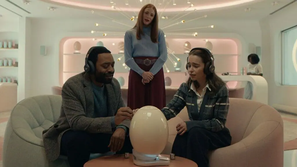 Chiwetel Ejiofor and Emilia Clarke sit in a futuristic office wear headphones as they listen to their new baby while its growing inside a large artificial egg pod in the sci-fi satire film THE POD GENERATION.