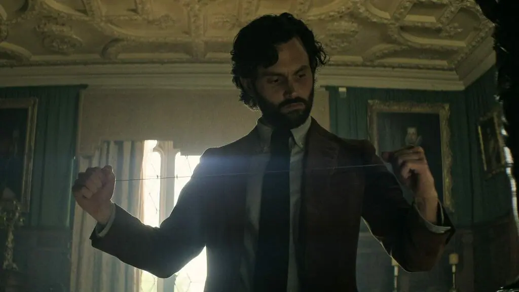 Penn Badgley as Joe Goldberg stretches out a thin piano wire in a fancy London ballroom with classic paintings on the walls in YOU Season 4 Part 1 on Netflix.
