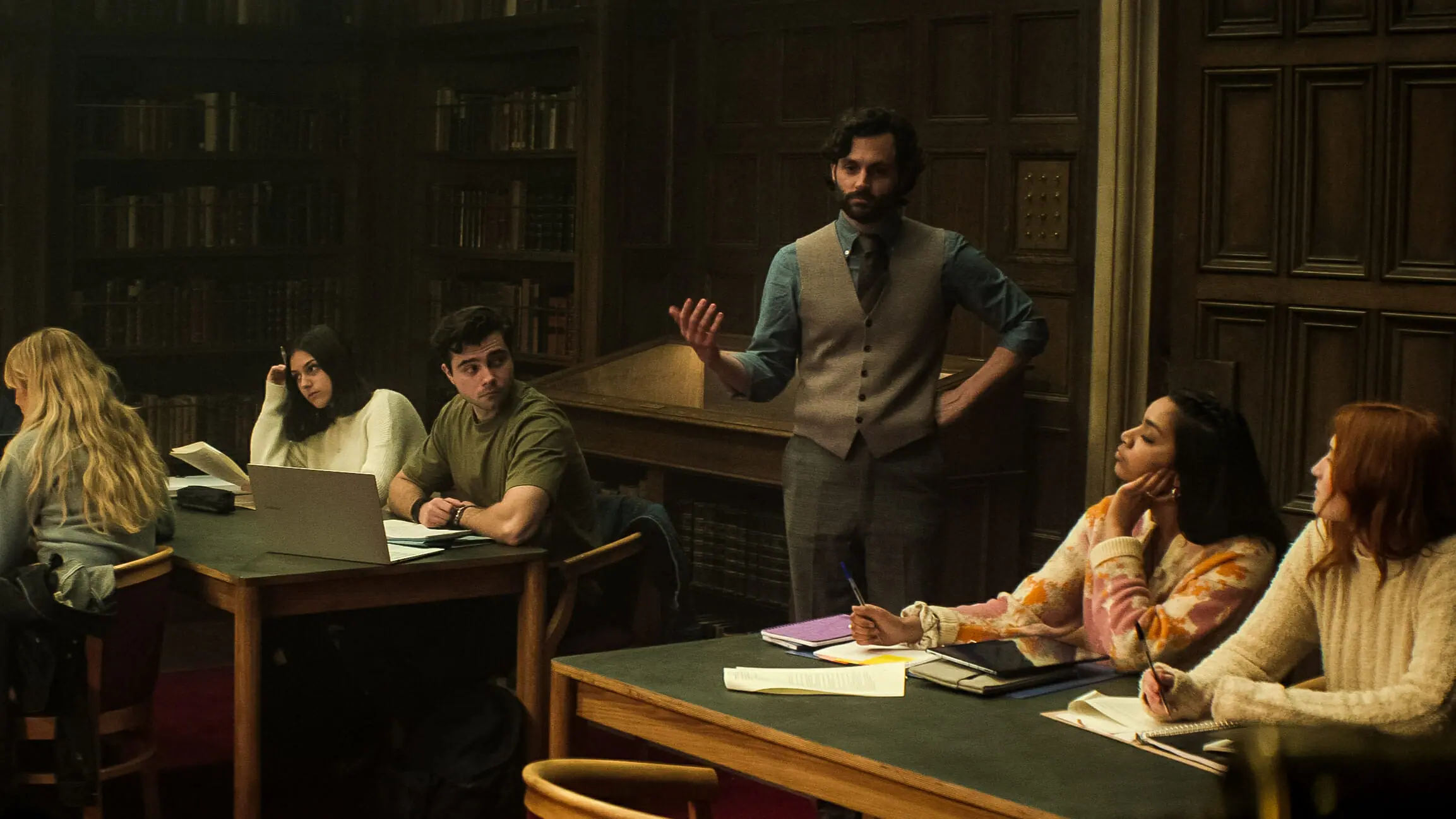 Penn Badgley as Joe Goldberg posing under his new identity professor Jonathan Moore while teaching a London Literature class full of young students in YOU Season 4 on Netflix.  