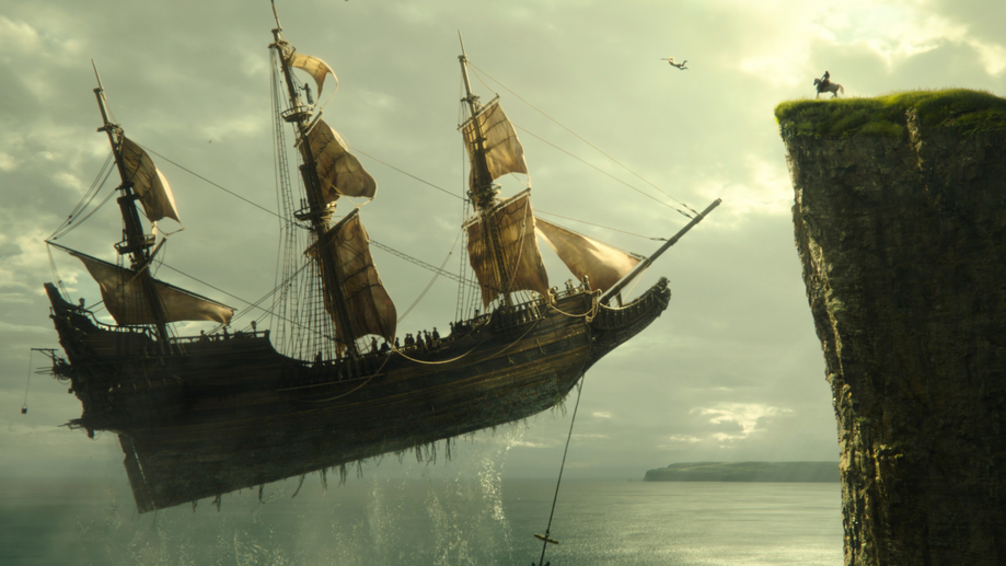 Captain Hook's pirate ship floats in the air next to a giant cliff with Peter Pan standing on top in the live-action Disney remake PETER PAN & WENDY coming to Disney+ in April 2023.