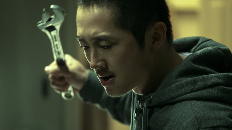 Steven Yeun stars as David Cho holding up a huge wrench and preparing to swing in the dark comedy thriller original series BEEF which is new on Netflix in April 2023.
