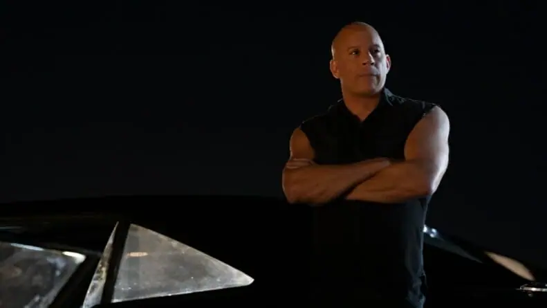 Vin Diesel stars as Dominic Toretto wearing a black vest and posing with his arms crossed while leaning on his new black charger in FAST X.
