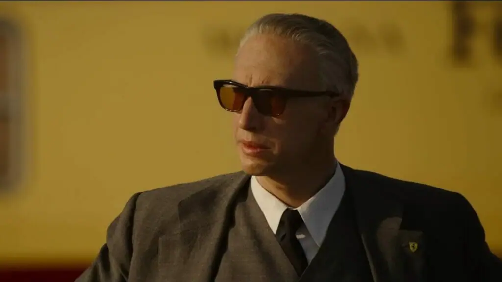Adam Driver stars as Enzo Ferrari posing in a gray suit with sunglasses while watching a car race in the biopic FERRARI directed by Michael Mann.