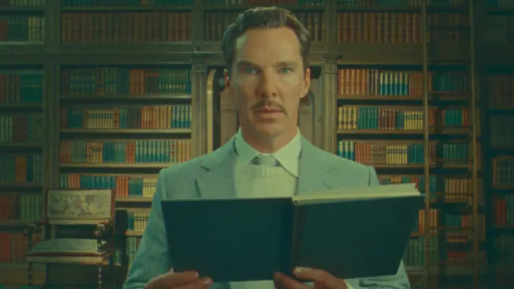 Benedict Cumberbatch as Henry Sugar breaks the fourth wall and stares into the camera as he opens up a blue book in a colorful library in THE WONDERFUL STORY OF HENRY SUGAR on Netflix.