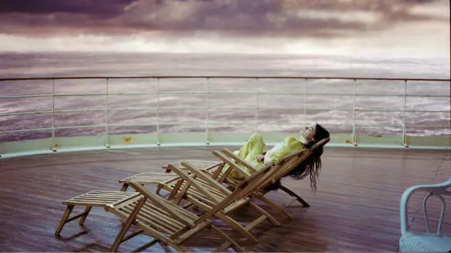 Emma Stone stars as Bella Baxter laying on the deck of ship in a beautiful yellow dress in front of an open violet-colored ocean in a cinematic shot from the movie POOR THINGS. 