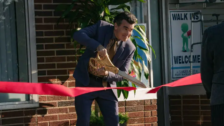 Brian d'Arcy James wears a fancy suit and tie as he cuts a large red ribbon with large scissors during the opening ceremony of a new pharmaceutical office building in the Netflix original film PAIN HUSTLERS.