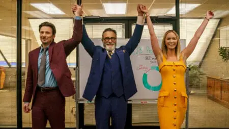 Andy Garcia raises the hands of Chris Evans and Emily Blunt in fancy suits and dresses during a celebration in their pharmaceutical sales office in the Netflix original movie PAIN HUSTLERS.