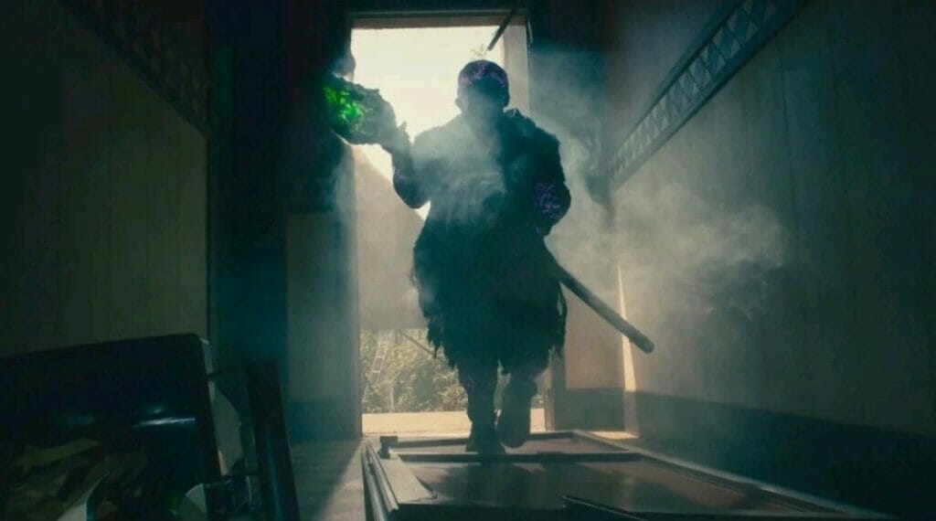 A silhouette of Peter Dinklage as the new Toxie entering a room with his green-glowing radioactive broom ready for battle in the 2023 THE TOXIC AVENGER remake.