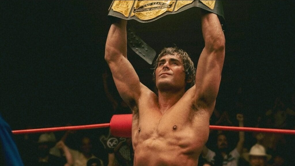 Zac Efron stars as the famous wrestler Kevin Von Erich holding up a golden championship title belt in the middle of the wresting ring in the A24 movie THE IRON CLAW.