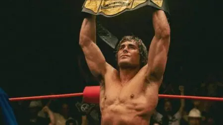 Zac Efron stars as the famous wrestler Kevin Von Erich holding up a golden championship title belt in the middle of the wresting ring in the A24 movie THE IRON CLAW.