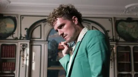 Josh Hutcherson gives a menacing stare as the villainous tech bro Derek Danforth while wearing a turquoise suit and a holding a gun over his shoulder in the action revenge thriller film THE BEEKEEPER.