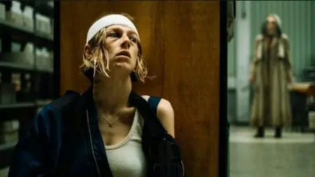 Hunter Schafer hides from a mysterious women behind a large shelf in an office building while wearing a head bandage in the horror film CUCKOO.
