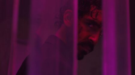 Dev Patel stars as the protagonist Kid drenched under neon purple lights and showing battle wounds on his angry face in the action revenge thriller film MONKEY MAN.