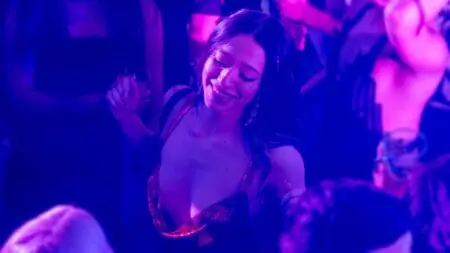 Mikey Madison wearing a beautiful red dress and a huge smile on her face dances in the middle of a packed Brooklyn night club with neon purple lighting in the film ANORA.