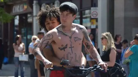 Barry Keoghan rides an electric scooter around the small English town of Dartford showing off his body fully of tattoos shirtless while his young daughter played by Nykiya Adams holds on to him from behind in the movie BIRD.