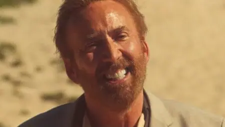 A close up of Nicolas Cage giving a huge eerie smile as he slowly loses his mind standing on a beach under the hot Australian sun in the psychological thriller THE SURFER.