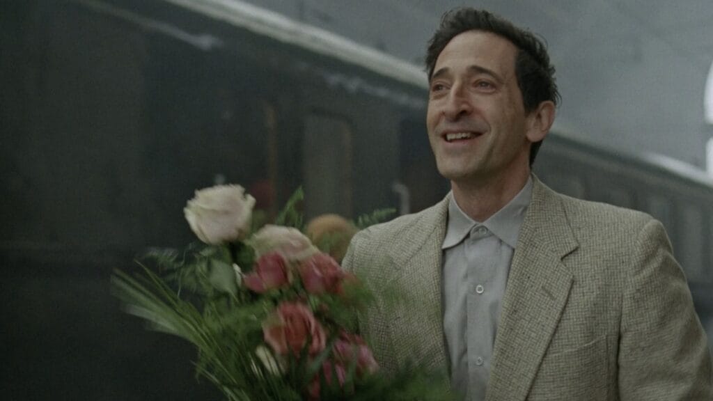 Adrien Brody holds a bouquet of flowers at a train station as he smiles and tries to hold back tears of happiness in the historical epic film THE BRUTALIST. 