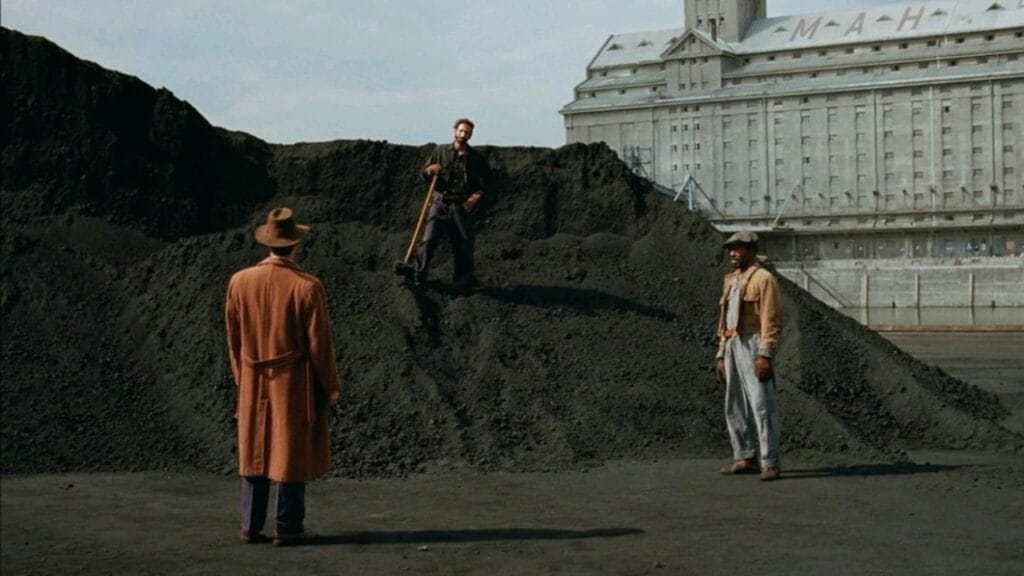 Adrien Brody stands on top of a giant dirt hill as he surveys the landscape of a huge construction site with two other colleagues looking by in the historical epic THE BRUTALIST.  