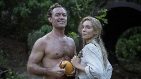 Jude Law gives a huge charming smirk as he peels a mango shirtless next to Vanessa Kirby as the two pose for a photograph in front of a jungle in the Galápagos Islands from the new thriller film EDEN.