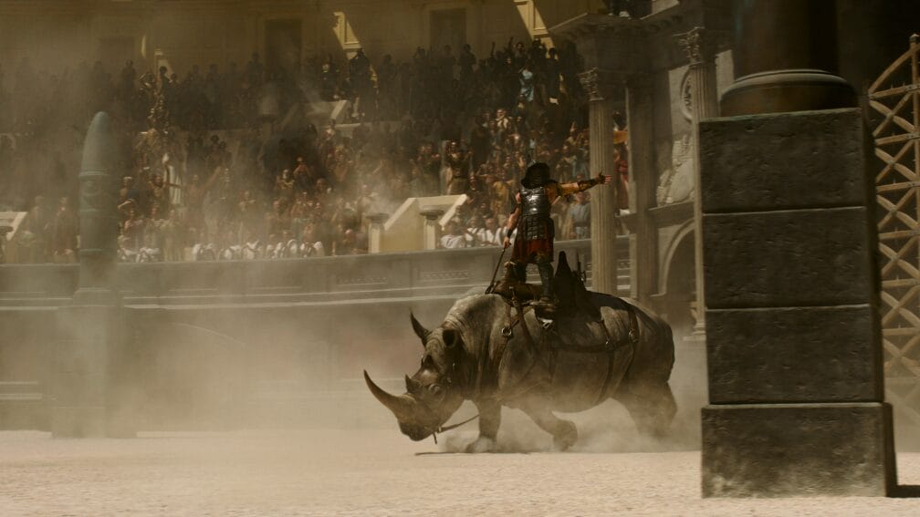 A beefy gladiator rides on top of a huge armored rhinoceros in the middle of the Colosseum as packed crowds watch from the stands in the legacy sequel GLADIATOR II.  